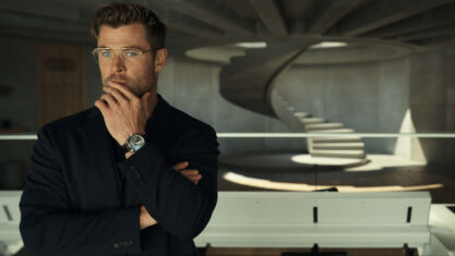 Chris Hemsworth as Steve Abnesti in Netflix's Spiderhead. He's dressed in a black suit and is wearing glasses. He has one hand one his chin as he stares forward contemplatively