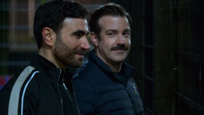 Ted Lasso, played by Jason Sudeikis, watches Roy Kent, played by Brett Goldstein. Both are smiling.