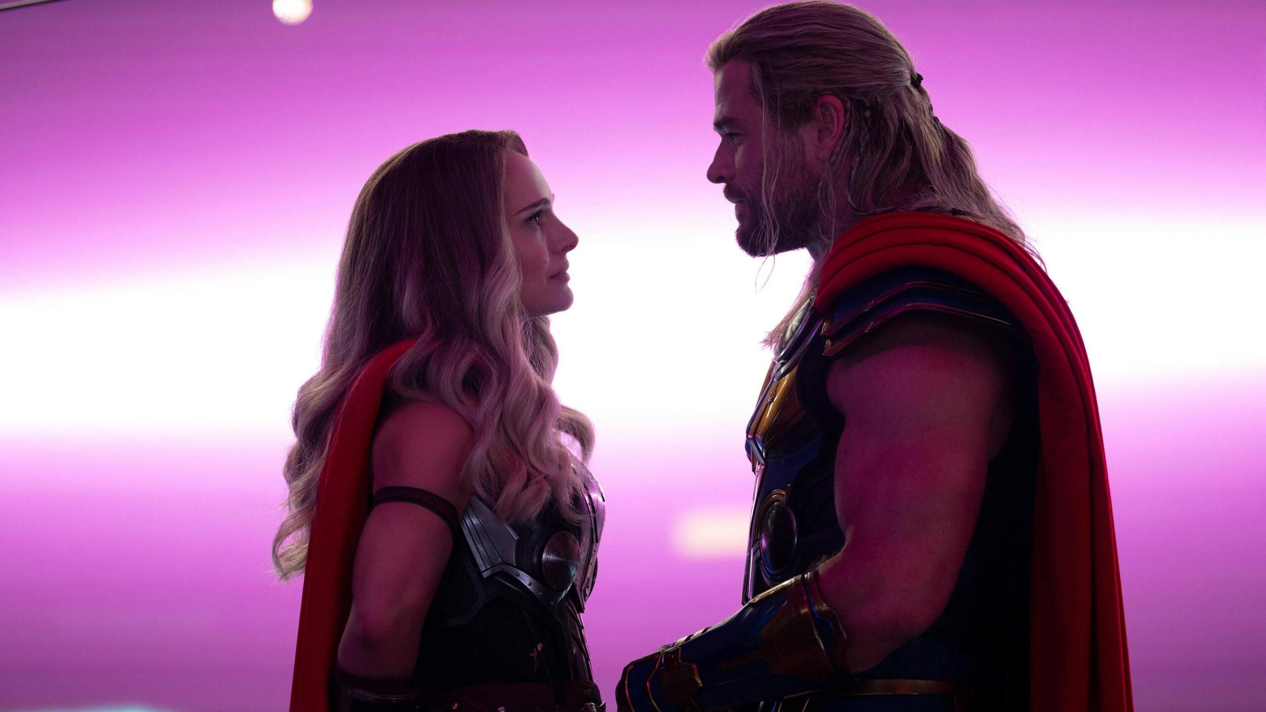 Natalie Portman as The Mighty Thor and Chris Hemsworth as Thor in Thor: Love and Thunder. The two are standing close to one another with a bright purple light behind them.