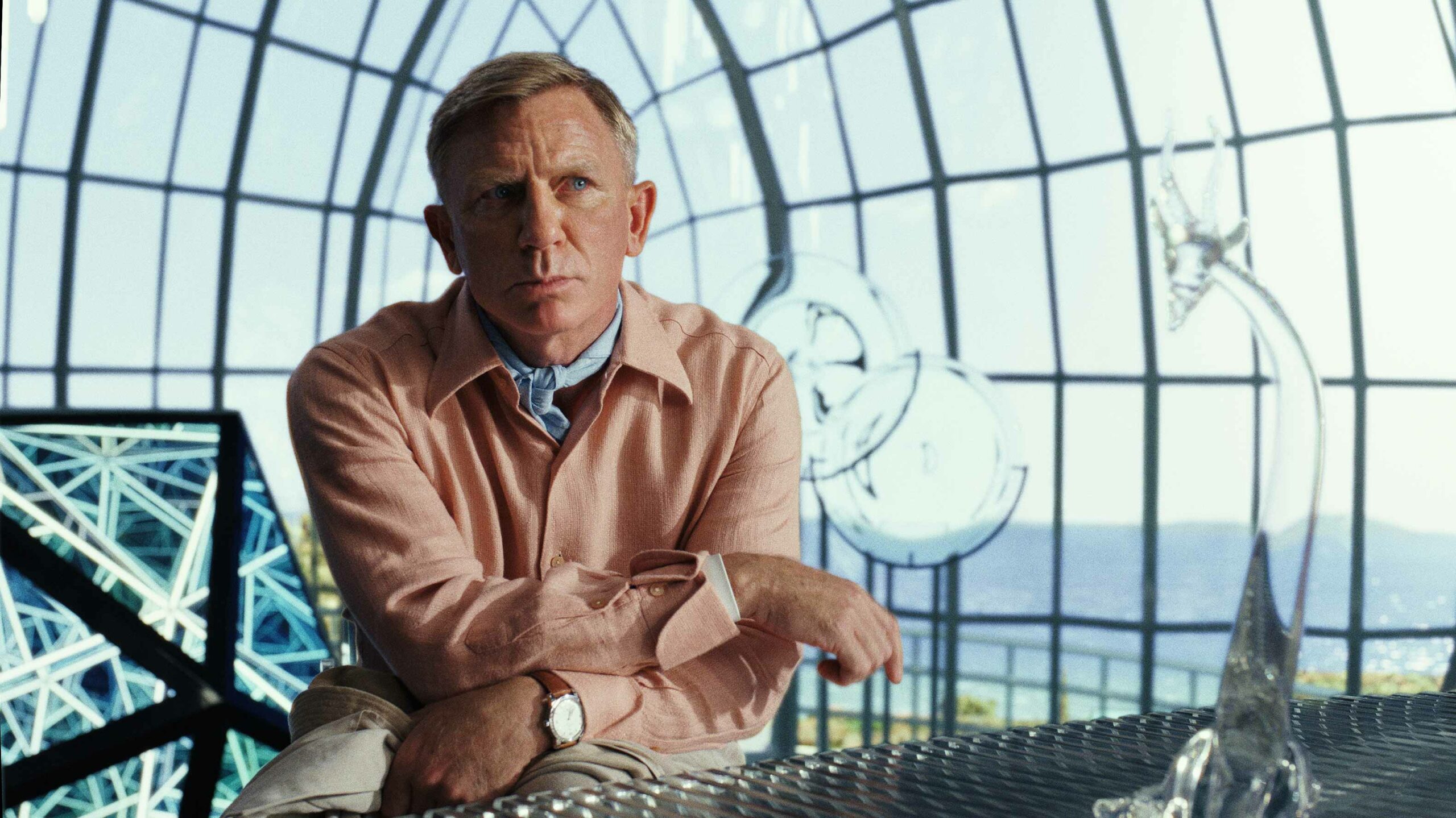 Daniel Craig as Benoit Blanc in Glass Onion: A Knives Out Mystery. He's wearing a pink dress shirt and sitting down while leaning forward. He looks deep in thought.